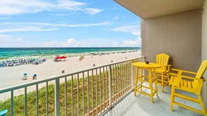 Un balcon avec des chaises jaunes et une table donnant sur une plage parsemée de parasols et fréquentée par des vacanciers.