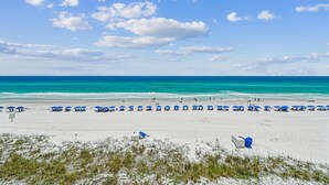 A beach with white sand, clear blue water, and a few people. There are blue umbrellas and lounge chairs along the shore. - Destin Gulfgate 2 Bedroom, 2 Bath Gulf-Front Condo! (Destin)