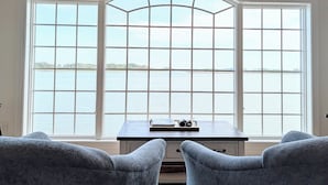 A room with a large arched window overlooking a body of water, a desk with a laptop and binoculars, and two grey armchairs. - Luxurious 5BR Inlet View Home with Elevator – South Shore Marina (North Bethany)