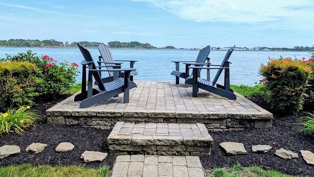 Two Adirondack chairs facing a body of water with a stone-paved deck and a stone border. - Luxurious 5BR Inlet View Home with Elevator – South Shore Marina (North Bethany)