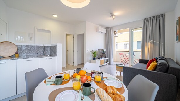 Una sala de estar moderna con un sofá gris, una mesa de comedor dispuesta para el desayuno y una zona de cocina con encimera de granito.