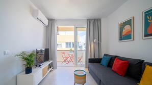 Una sala de estar moderna con televisor de pantalla plana, sofá gris, cojines coloridos, una mesita auxiliar y una planta en maceta.