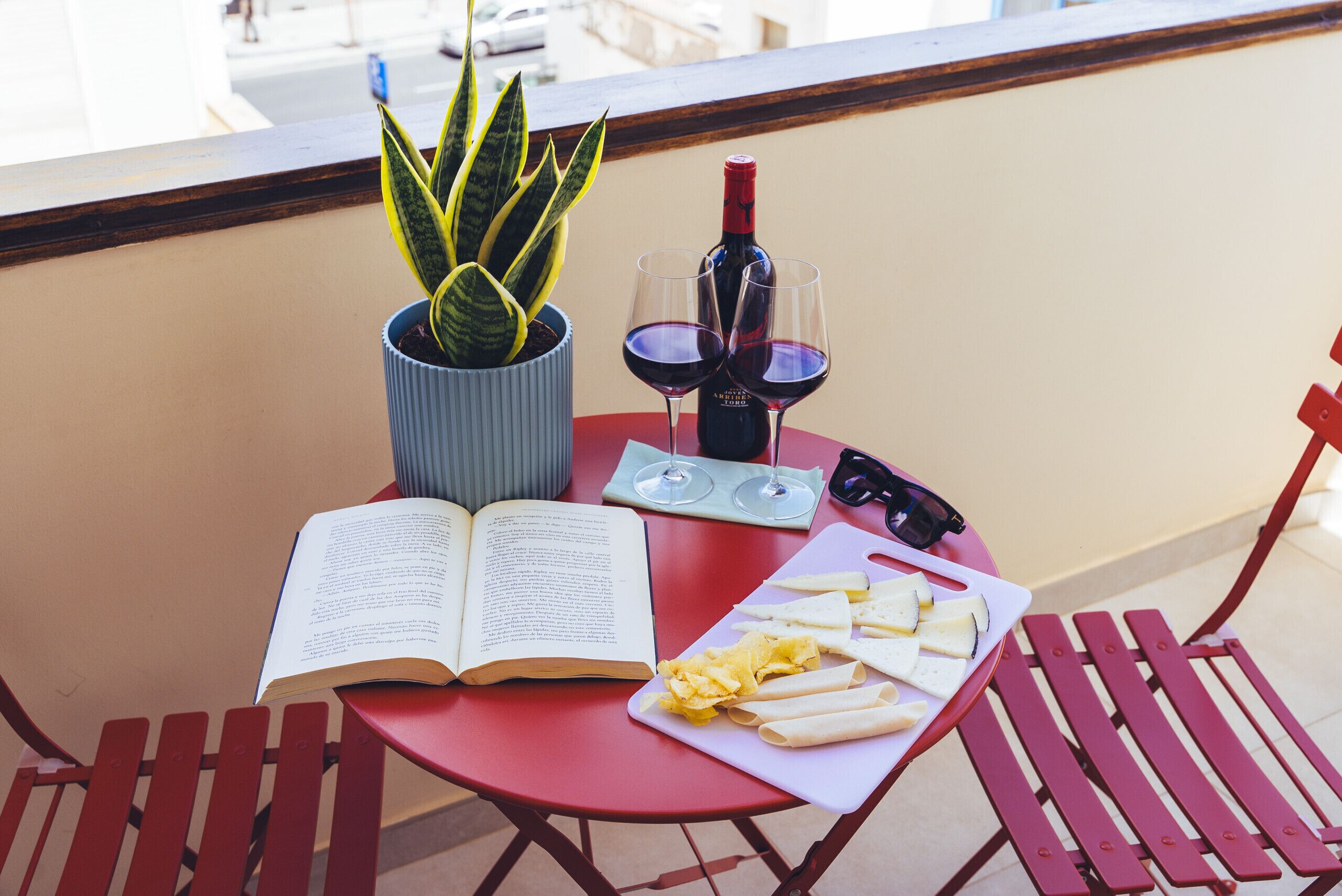 Une table rouge avec un livre, une bouteille de vin, des verres, une plante et du fromage, le tout sur un balcon.