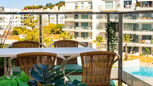 Un balcon avec des fauteuils en osier et une table, donnant sur une piscine et des bâtiments modernes.
