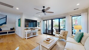 Ampla sala de estar com piso de madeira, ventilador de teto, televisão, sofá, mesa de centro e porta de vidro deslizante para um pátio externo.