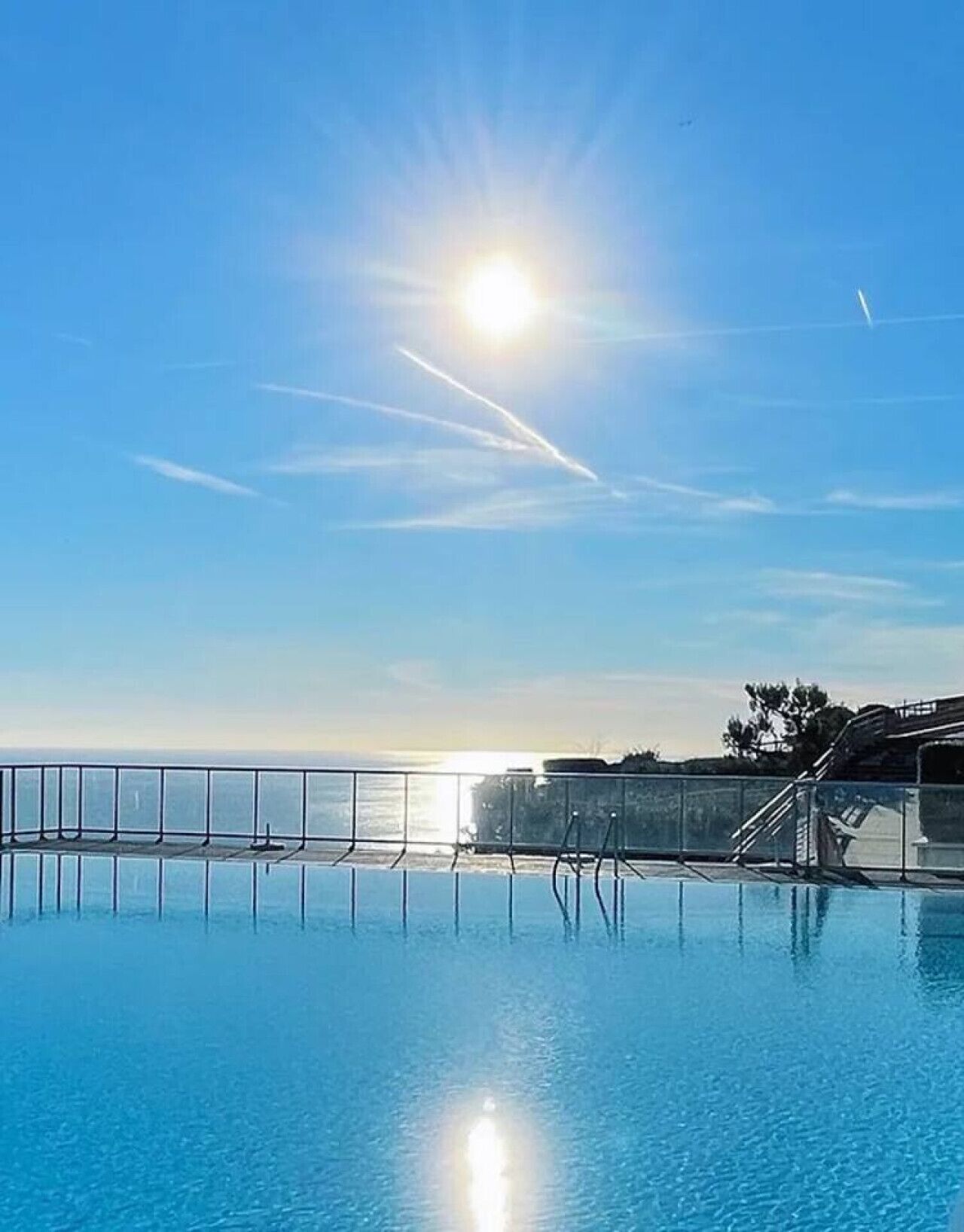 Un ciel d’un bleu limpide, avec le soleil qui brille sur une piscine, offrant une vue sur l’océan et un bâtiment en arrière-plan.