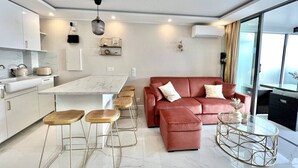 Una sala de estar moderna con un sofá rojo, una mesa de centro de vidrio y una cocina con gabinetes blancos y encimera de mármol.