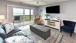 Sala de estar com sofá de couro, repouso para os pés e tapete estampado. Há uma televisão na parede, uma poltrona e uma mesinha com objetos decorativos.