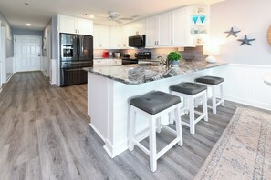 A modern kitchen with white cabinetry, a stainless steel refrigerator, a built-in oven, and a granite countertop. There are three bar stools and a small island with a plant. - Oceanfront 3 BR  Condo with a Shared Pool! Sea Shadow 103   (Surfside Beach)