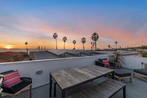 A rooftop terrace with a sunset view, featuring a wooden table and chairs, and several palm trees. - Ocean View Gem with Rooftop Deck and Hot Tub (San Diego)