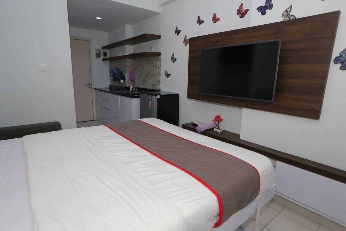 A bedroom with a bed, a TV mounted on the wall, and a kitchenette in the background.