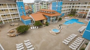 A hotel courtyard with multiple swimming pools, lounge chairs, and a central building with a tiled roof. - Book Lazy Waves 414B | Sandbridge Vacation Condo (Virginia Beach)