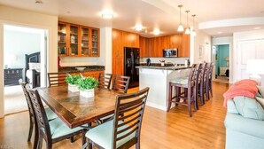 A modern kitchen with wooden cabinets, a central island, and a dining area with a wooden table and chairs. - Book Lazy Waves 414B | Sandbridge Vacation Condo (Virginia Beach)