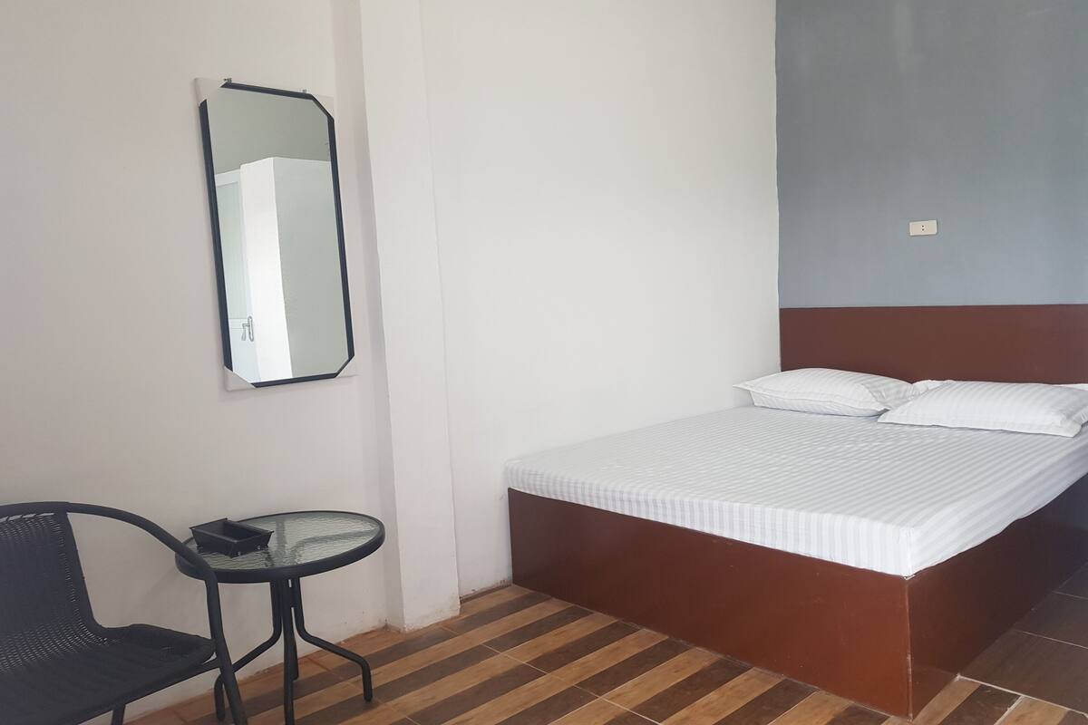 Een slaapkamer met een bed, een klein tafeltje, een stoel en een spiegel aan de muur.