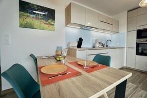 A modern dining area with a wooden table set for two, a kitchen in the background, and a painting on the wall.