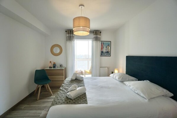 A hotel room with a bed, a desk, a chair, a round mirror, and a window with curtains.