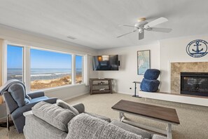 Una sala de estar con un ventanal grande que da a la playa, un televisor de pantalla plana, chimenea y ventilador de techo.