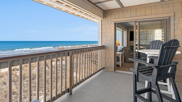 Een balkon met uitzicht op het strand en de oceaan. Er staat een tafel met twee stoelen en een lamp.