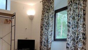 A room with a bed, a TV on a stand, a window with curtains, and a wooden floor. - Camera immersa nel verde (Sorrento)