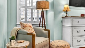 A room with a light blue wall, a white dresser with black handles, a round wooden table, a floor lamp, and a chair with a patterned cushion.