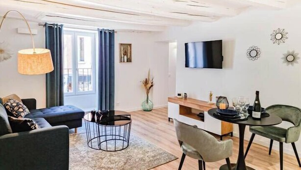 A modern living room with a sofa, a coffee table, a dining area with chairs, a TV mounted on the wall, and a large window with curtains. - ELEGANCE in the heart of the old town 4 **** (Annecy)