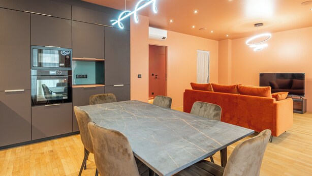 A modern kitchen with built-in appliances, a dining area with a marble table, and a living room with a sofa and TV.