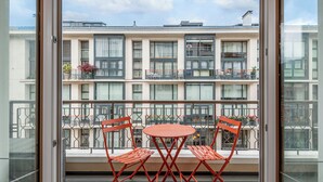 A balcony with two red chairs and a small table, overlooking a multi-story building with balconies and windows.