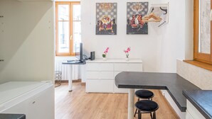 Una cocina moderna con encimera blanca, dos taburetes negros y un armario blanco. Hay dos cuadros en la pared, una ventana con persianas de madera y una puerta que da al exterior.
