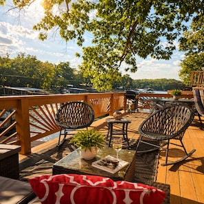 A wooden deck with wicker chairs, a table with a plant and a glass, and a view of a lake. - Granary House – Lakefront Bliss on Sunrise Beach (Osage Township)