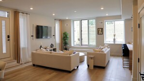 A modern living room with a sofa, armchairs, a coffee table, and a TV. There is a large window with curtains, a plant, and a sliding door leading to a balcony.