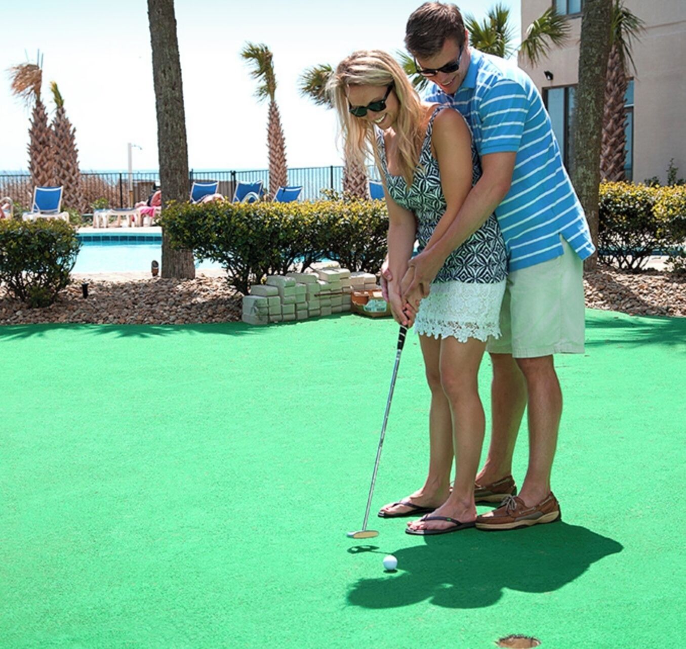 A couple playing golf on a green field with a pool and palm trees in the background.