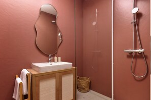 A bathroom with a wooden vanity, a white sink, a mirror, a shower with a handheld showerhead, and a woven basket on the floor.