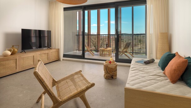 Un salon moderne doté d’une grande porte-fenêtre coulissante donnant sur un balcon, d’un téléviseur à écran plat posé sur une console en bois, d’un fauteuil en osier et d’un canapé agrémenté de coussins.