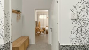 Un couloir avec un banc en bois, une petite étagère avec des plantes et une porte donnant sur une pièce où se trouvent un lit et une chaise.