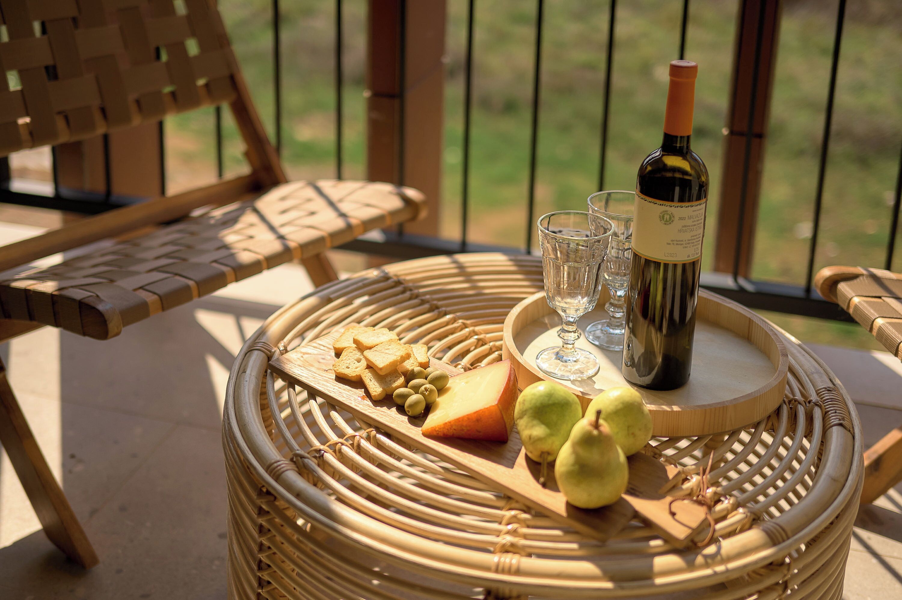 A wicker table with a bottle of wine, glasses, cheese, and pears on a balcony.