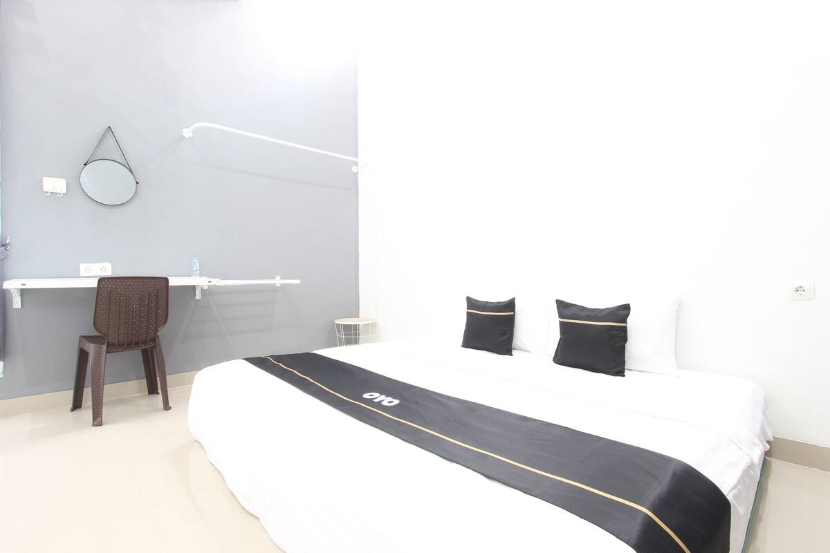 Quarto de hotel com cama, poltrona, escrivaninha, espelho e luminária fixada na parede.
