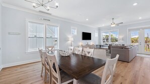 A spacious dining area with a dark wooden table and white chairs, a modern kitchen with a flat-screen TV, and a view of the beach through large windows. - Pet-Friendly Retreat with Hot Tub and Views (Port St. Joe)