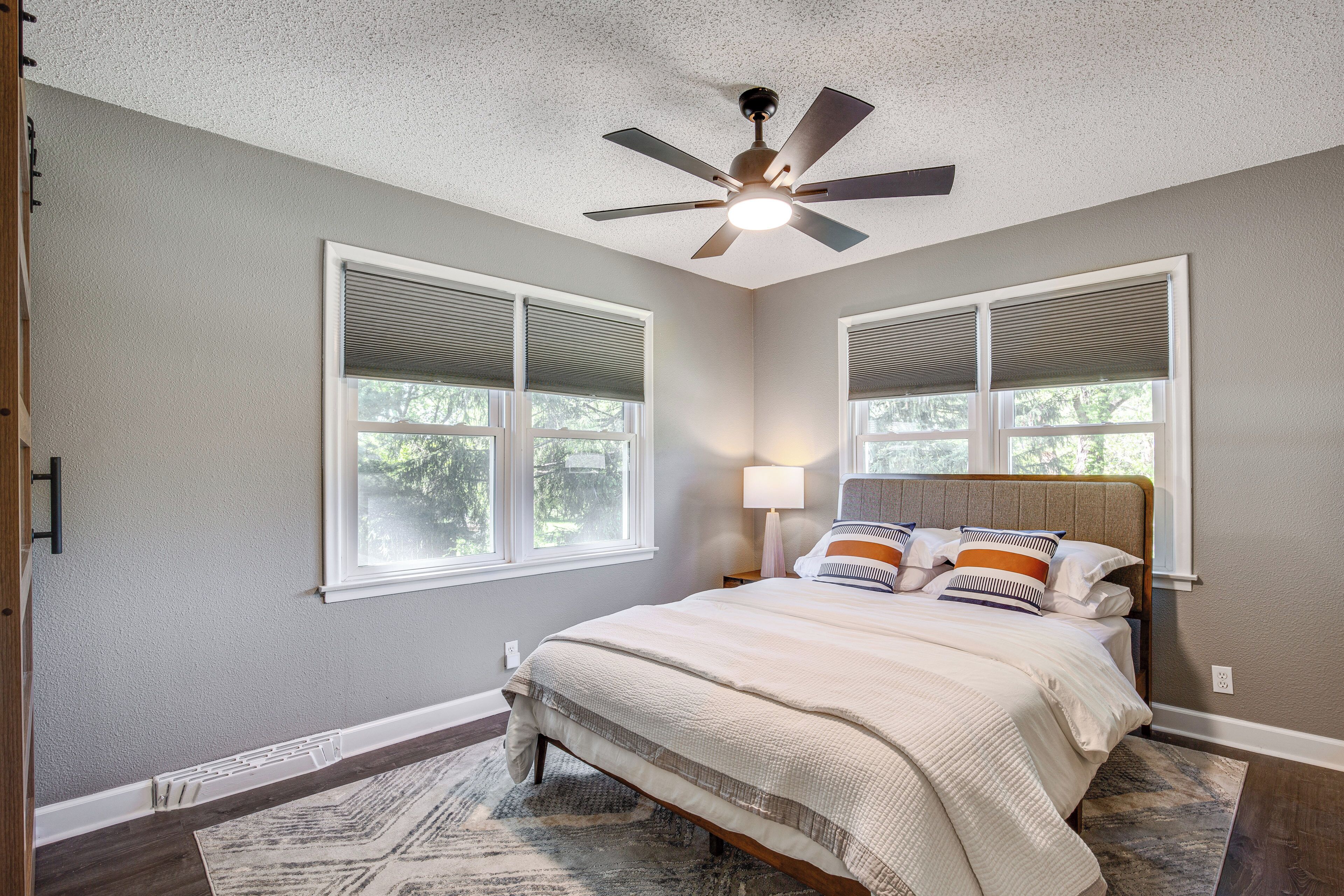 A bedroom with a bed, a ceiling fan, two windows with blinds, a nightstand with a lamp, and a patterned rug.