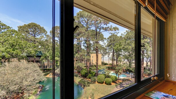 A view from a window of a room with a wooden floor and a magazine on it. Outside, there is a pool surrounded by trees and a building.