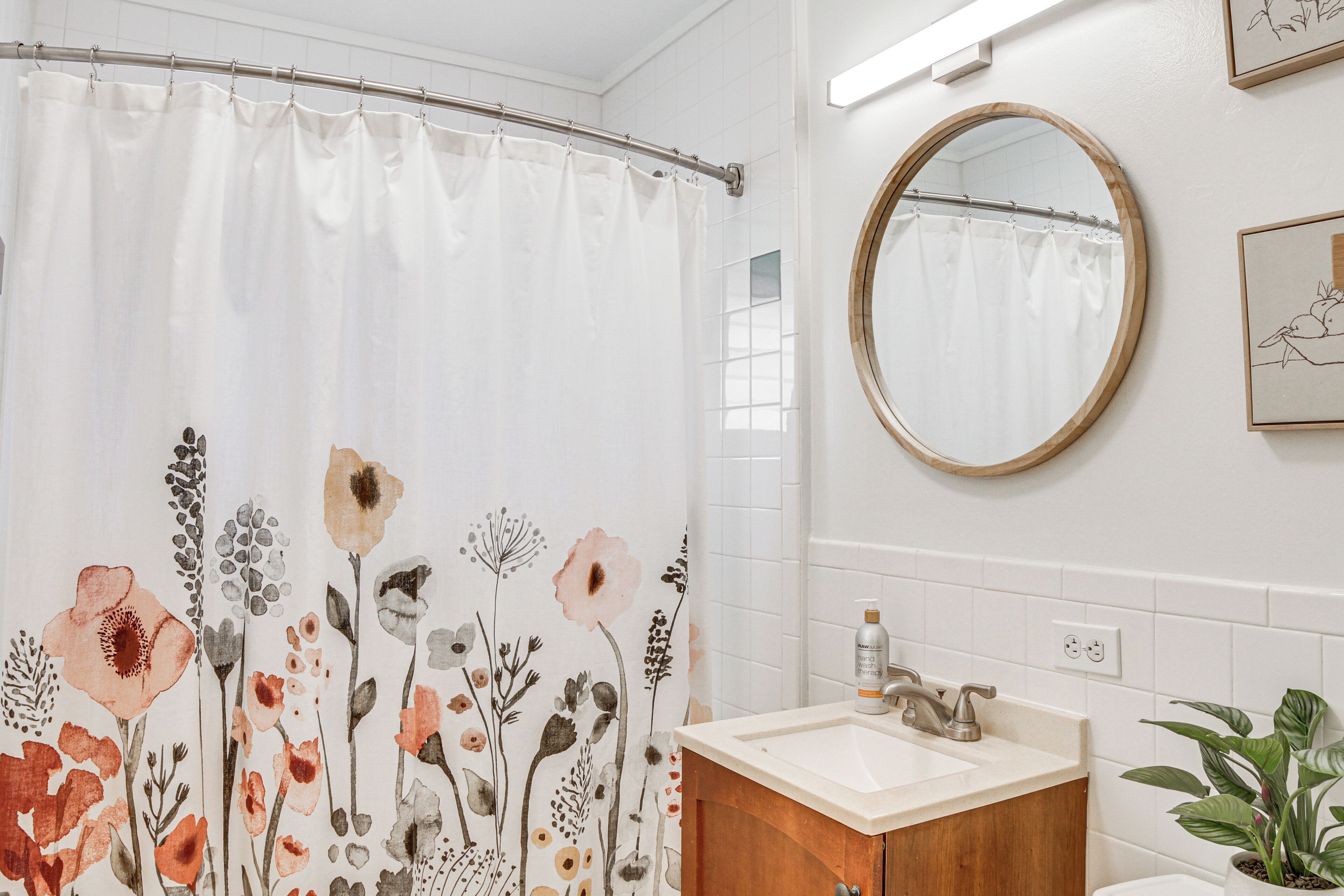 A bathroom with a shower curtain featuring floral patterns, a round mirror, a sink with a wooden cabinet, and a potted plant.