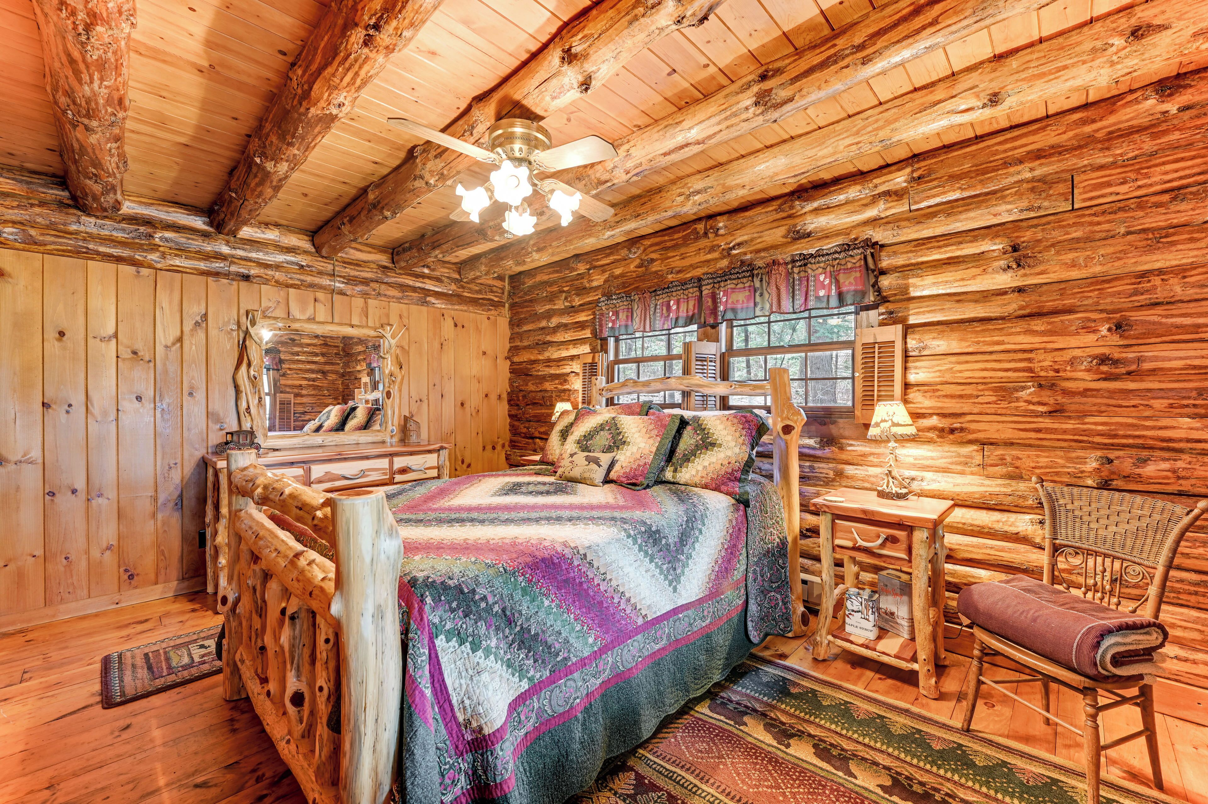 A log cabin bedroom with a bed, wooden walls, a ceiling fan, and a window with shutters.