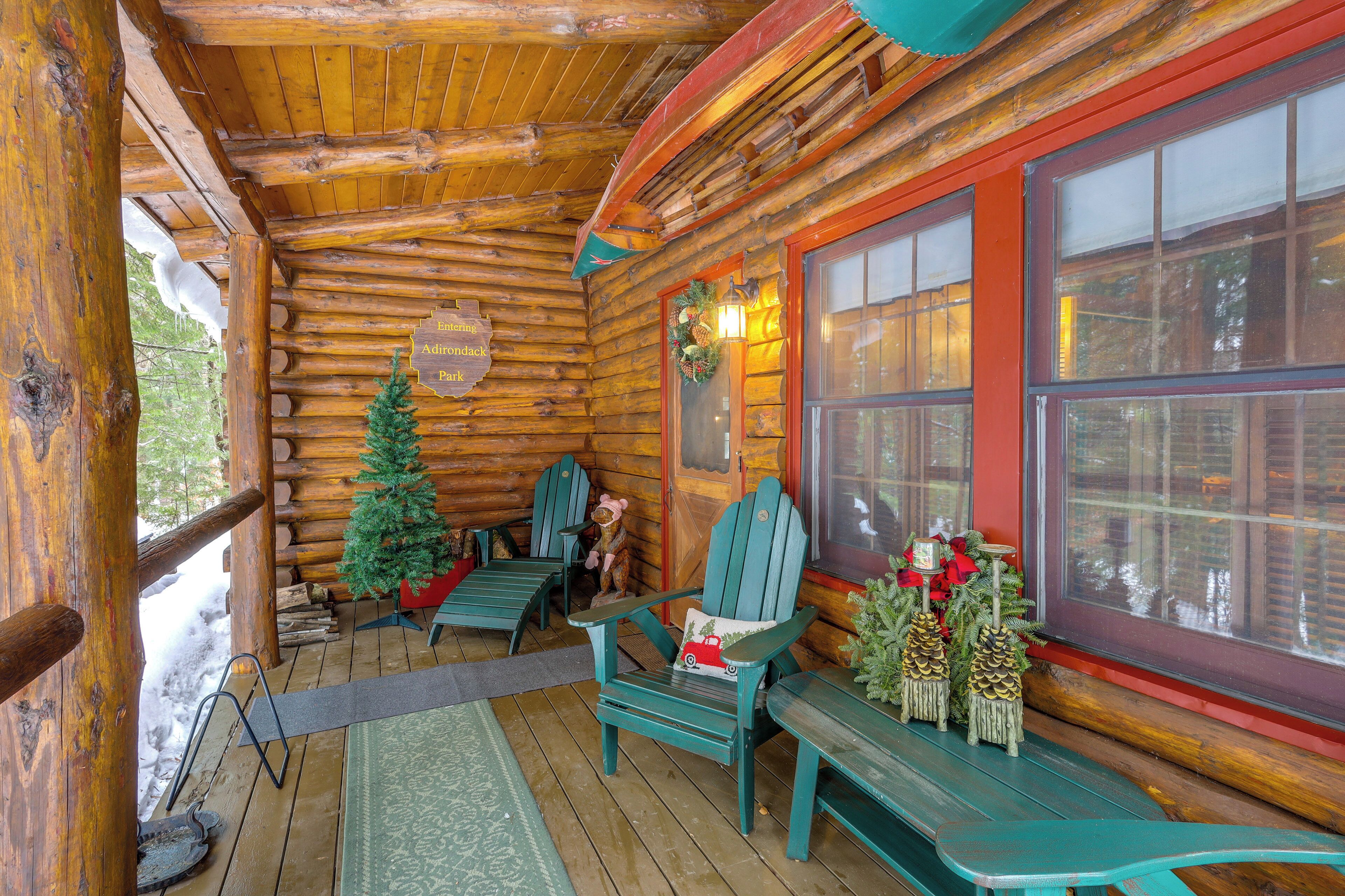 En timmerstuga med en veranda av trä, gröna Adirondack-stolar, en bänk och ett julgran.
