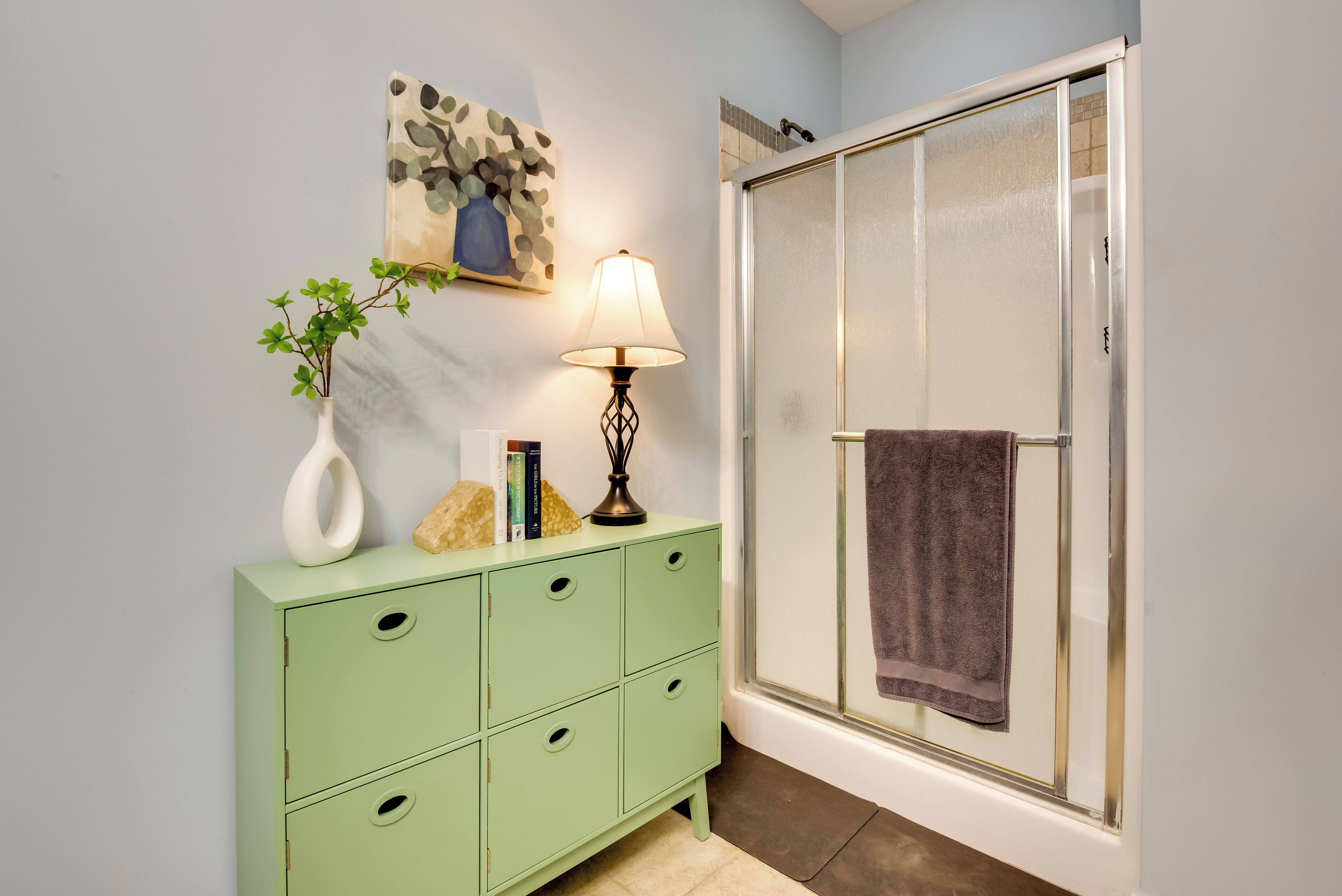 A bathroom with a green vanity, a shower with a glass door, a towel hanging on the door, and a wall-mounted artwork.