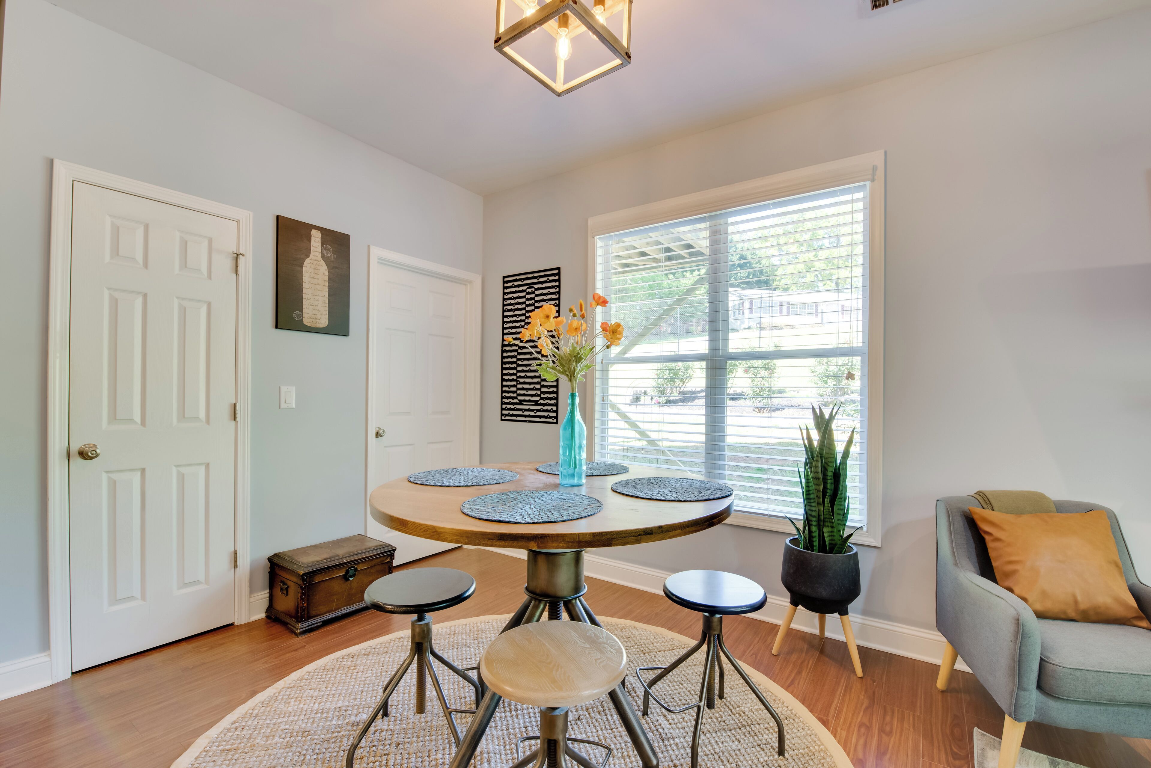 A dining area with a round table, four stools, a vase with flowers, a potted plant, and a chair with a cushion.