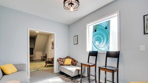 Ein modernes Wohnzimmer mit einem grauen Sofa, gelben Kissen, einem gemusterten Teppich und einem großen Fenster mit einem blauen spiralförmigen Kunstwerk.