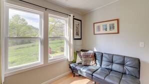 Ein Zimmer mit Ledersofa, zwei großen Fenstern mit Vorhängen, einem gerahmten Bild an der Wand und Blick ins Grüne.