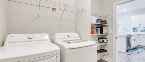 Un espace buanderie comprenant deux machines à laver et deux sèche-linge blancs, un étendoir et un coin cuisine avec un évier et des placards.