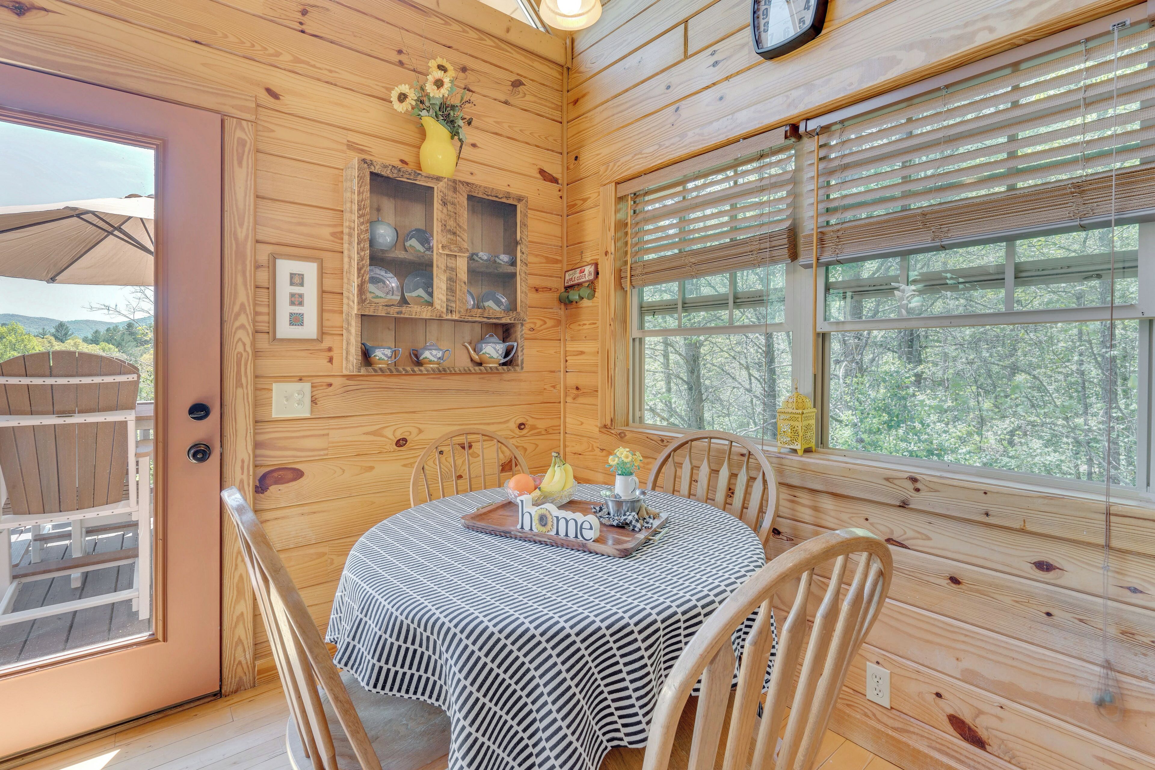 A wooden cabin interior with a dining table set for four, surrounded by wooden chairs. A wooden shelf with dishes and a vase of flowers is above the table.