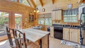 Interior de uma cabana de madeira com mesa de jantar, cadeiras, cozinha e grandes janelas.
