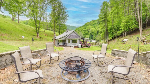 A rustic outdoor seating area with a fire pit, surrounded by chairs, in a lush green landscape with a house in the background. - Cozy Mars Hill Farmhouse w/ Scenic Views (Mars Hill)